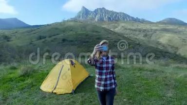 迷人的女孩徒步旅行使自拍在智能手机的背景山区。 一个女孩站在帐篷附近露营